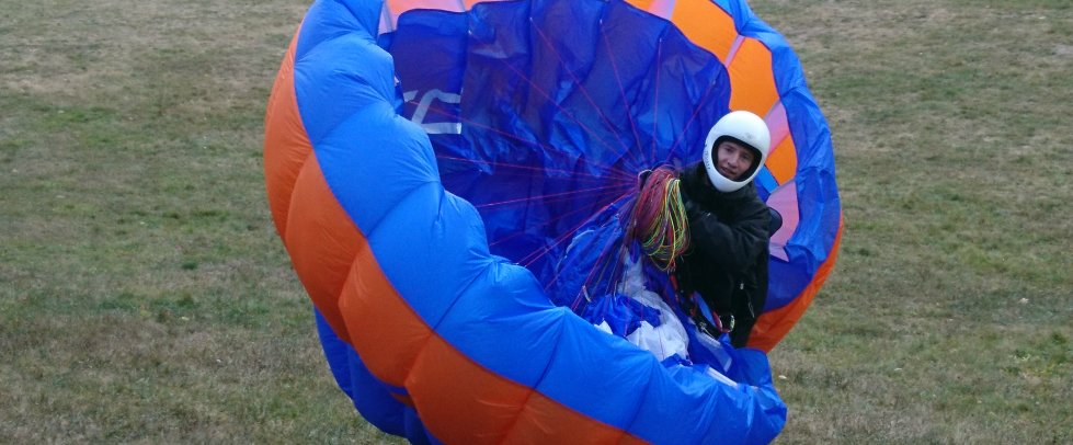 Behelmte Person zieht einen blau-orangen Gleitschirm auf einer Wiese auf