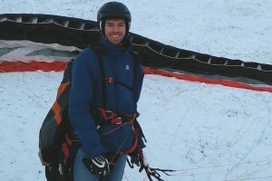 Bertram Hitthaler lächelnd in blauer Jacke mit Helm, Gleitschirm im verschneiten Hintergrund
