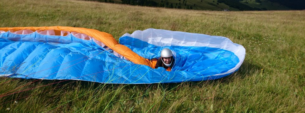 Auf einer Bergwiese ausgelegter blau-oranger Gleitschirm mit einer behelmten Person zwischen den geöffneten Zellen